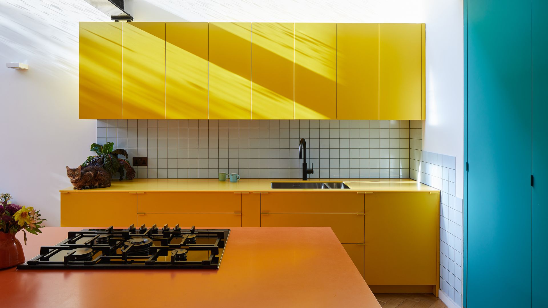 Cocina con los muebles amarillos, el antepecho de azulejos a cuadros, la isla naranja y la despensa azul