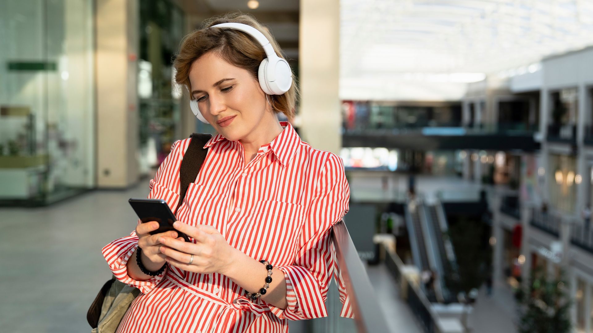 Mujer sonriendo en un centro comercial con unos auriculares y un teléfono móvil en la mano, con ropa de primavera-verano.