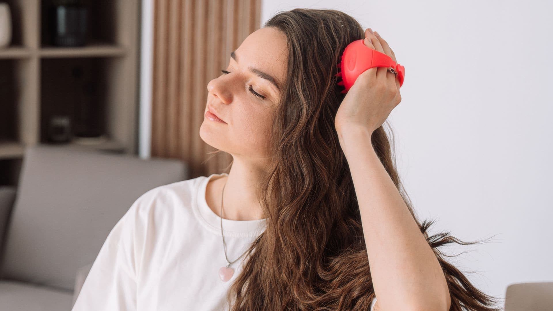 Mujer joven con pelo largo y ondulado, masajeándose el cuero cabelludo en su salón con un cepillo y los ojos cerrados