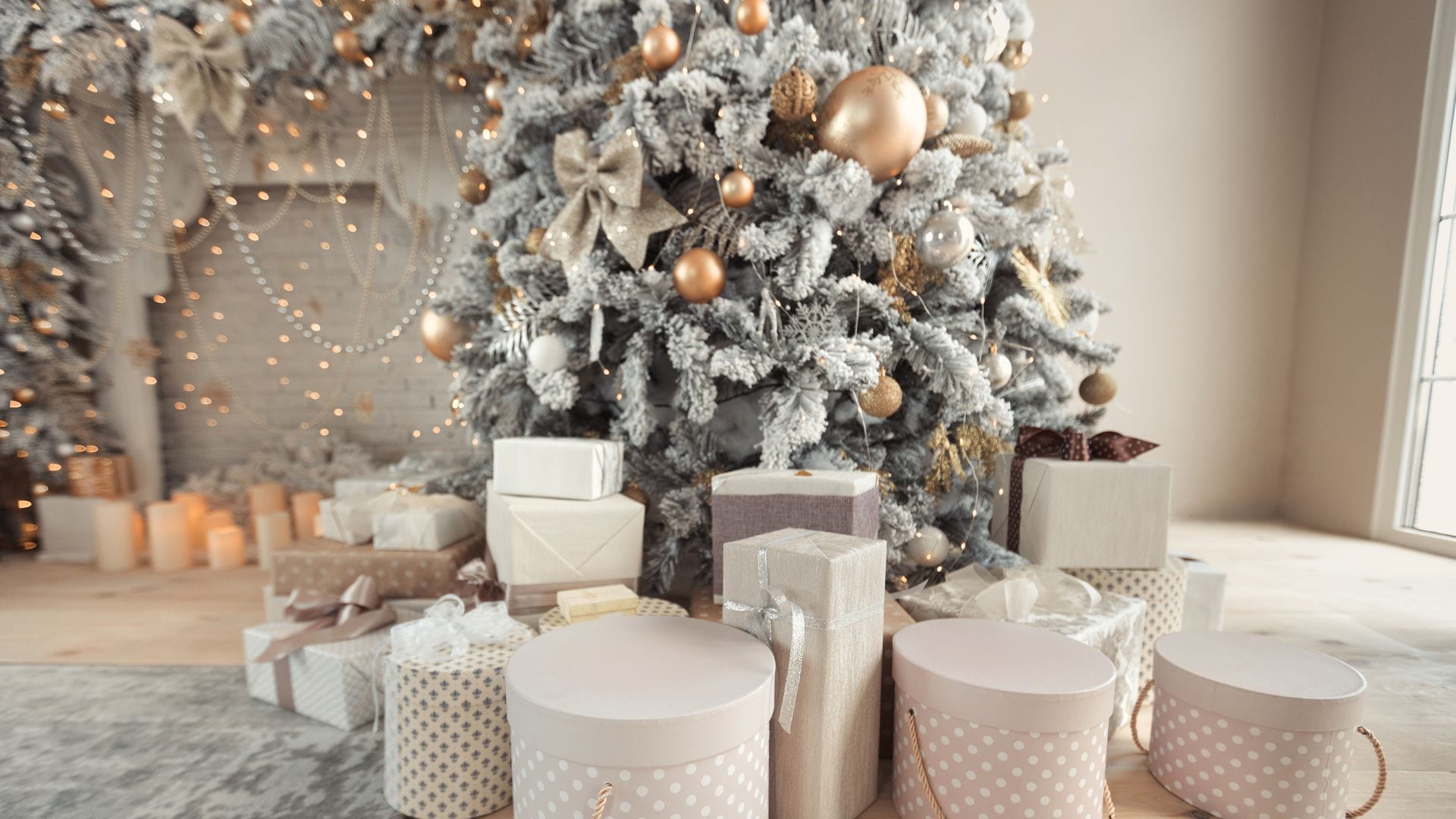 Interior de salón con chimenea, velas y un árbol de Navidad decorado con regalos. Gran árbol de nieve de diseño con luces y bolas en tonos beige, dorado y plateado.