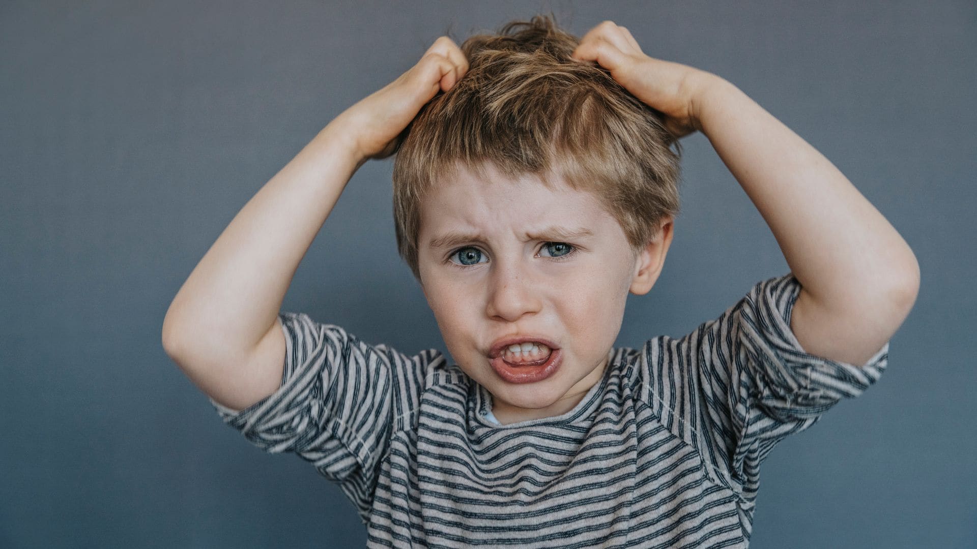 Niño pequeño mirando a la cámara rascándose la cabeza. Lleva camisetas de rayas azules y grises, es rubio y tienes los ojos azules.