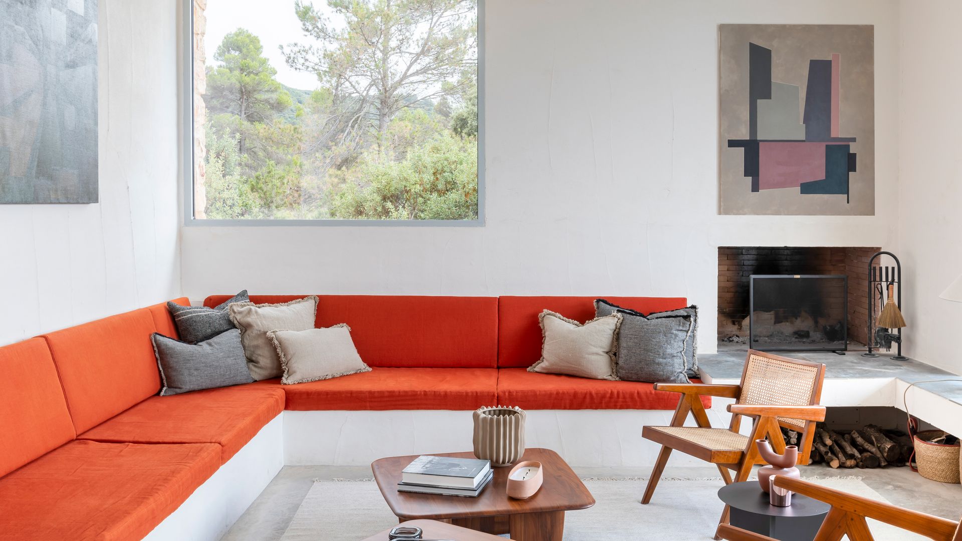 Salón con chimenea, sofá de obra con cojines rojos, sillas, mesa de centro de madera, cuadros en la pared, ventanal con vistas