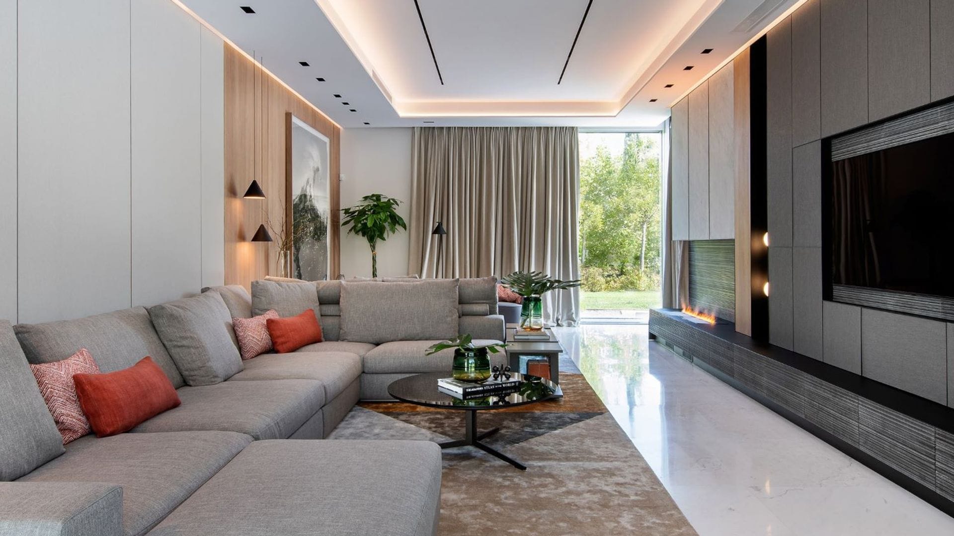 Salón con sofá gris XL, mueble de televisión de pared a pared, gris oscuro, chimenea, ventanal, iluminación en el techo, alfombra, mesa de centro, cortinas