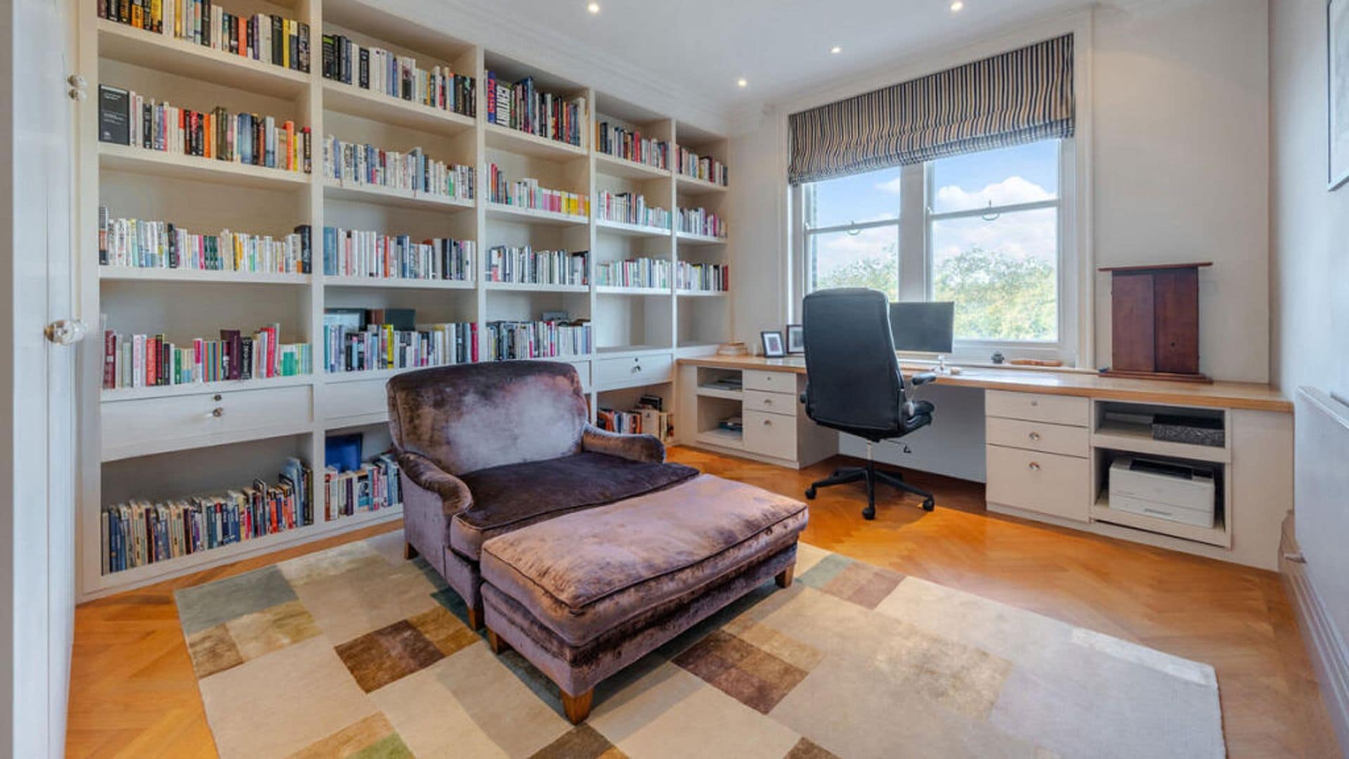 Despacho con librería de pared a pared, chaise longue, zona de escritorio bajo la ventana, silla de oficina y alfombra 