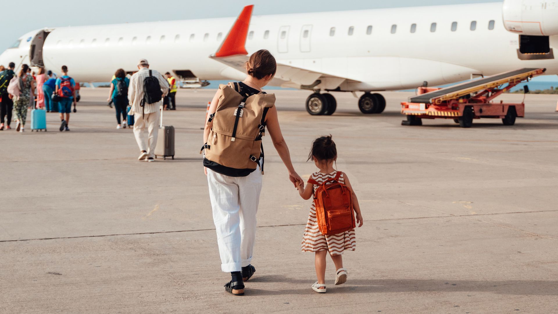 Madre e hija de espaldas caminando hacia un avión junto a otros pasajeros con mochilas en la espalda