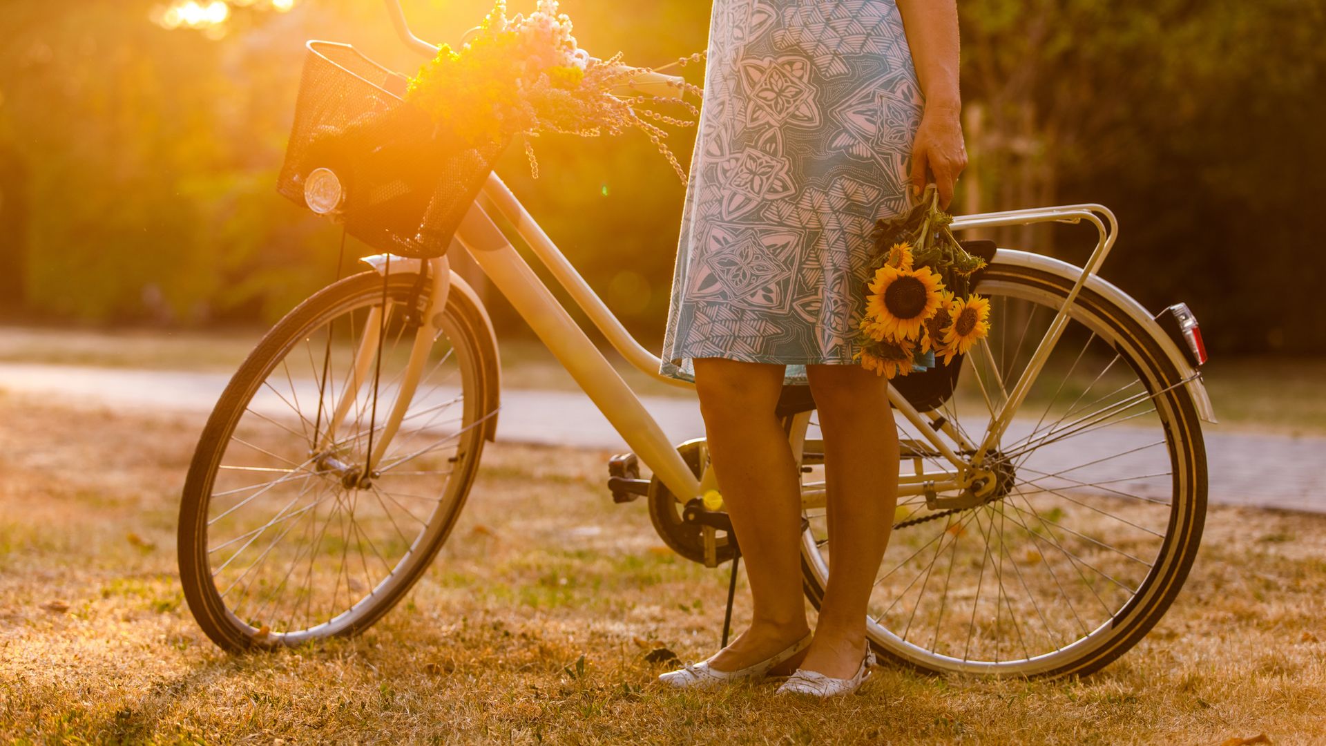 Mujer junto a una bicicleta en un parque con un ramo de girasoles en la mano, al atardecer. Lleva un vestido a la rodilla azul y solo se le ven las piernas.