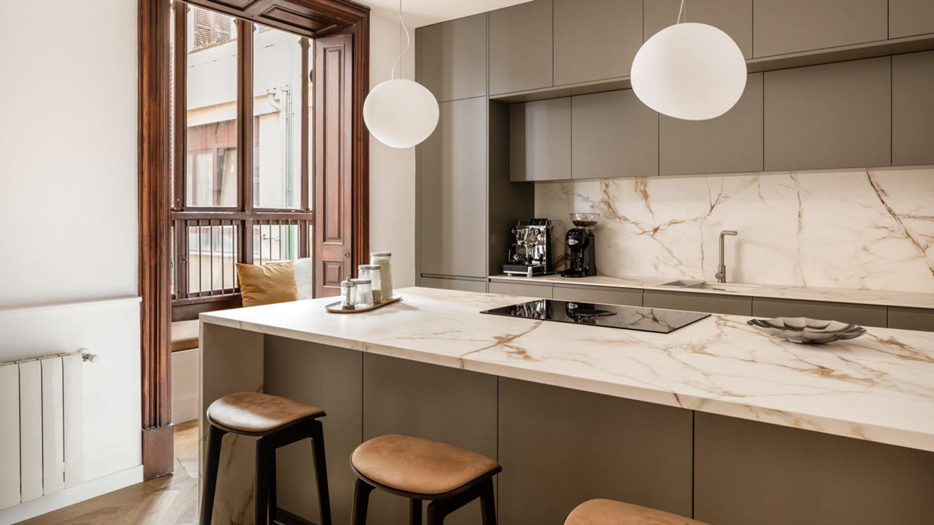 Cocina con muebles beiges, isla con encimera de mármol crema, ventana de madera, lámparas de techo blancas, taburetes