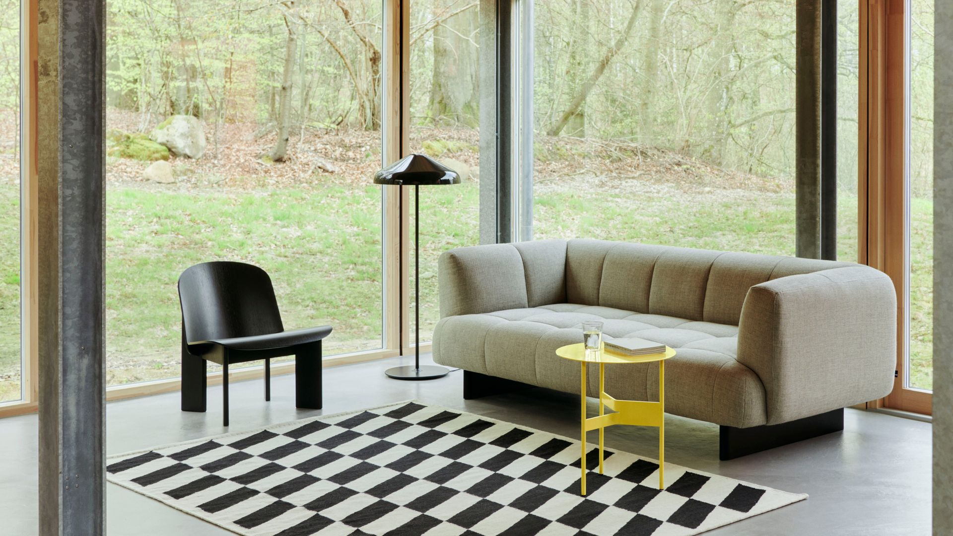 Salón con grandes ventanales con salida al jardín, sofá beige, mesa auxiliar amarilla, lámpara de pie negra, silla negra y alfombra damero negra y blanca