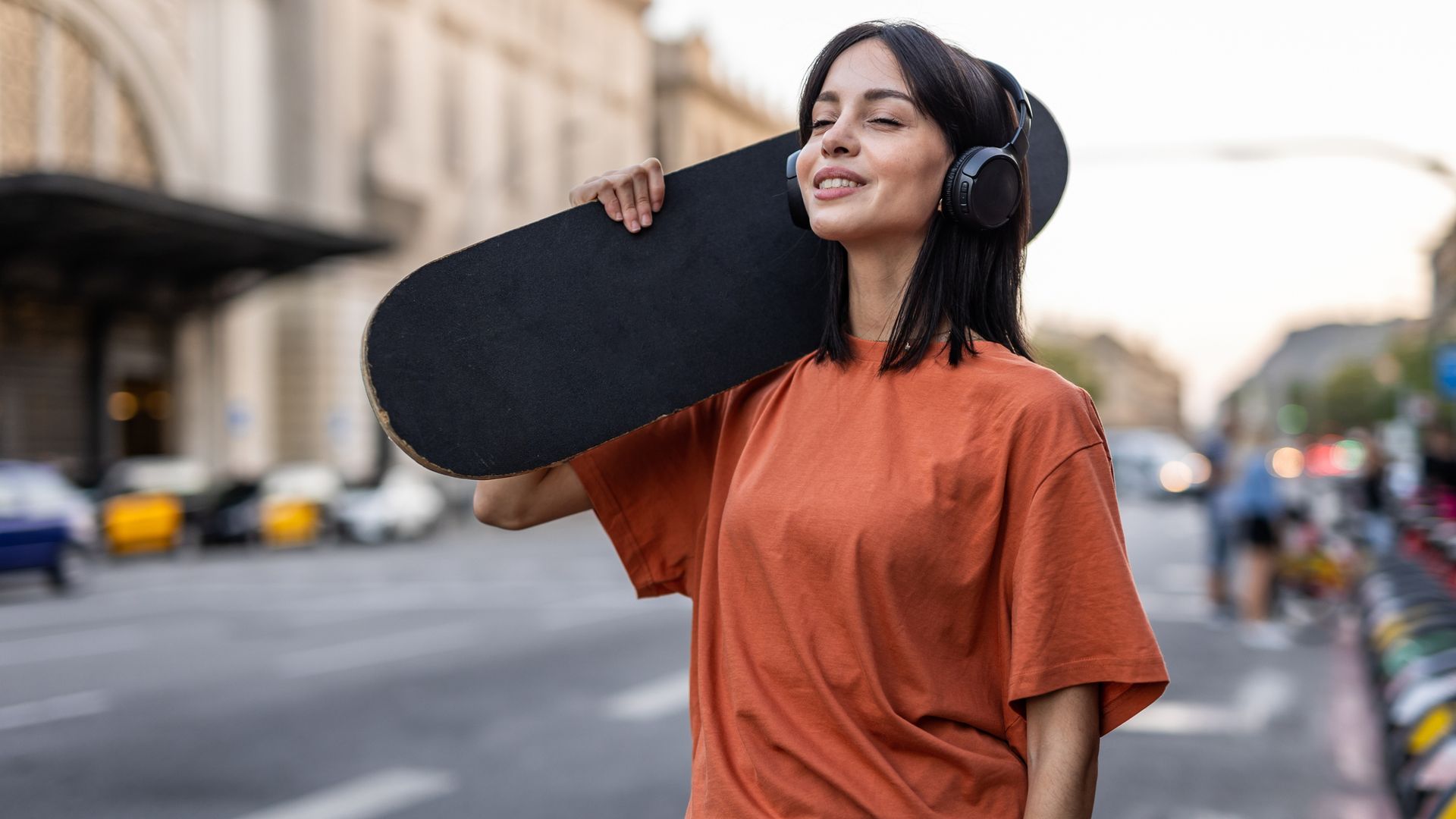 Mujer joven con auriculares inalámbricos sonriendo, en la calle con un patinete en los hombros. Ropa de otoño-invierno 2026.