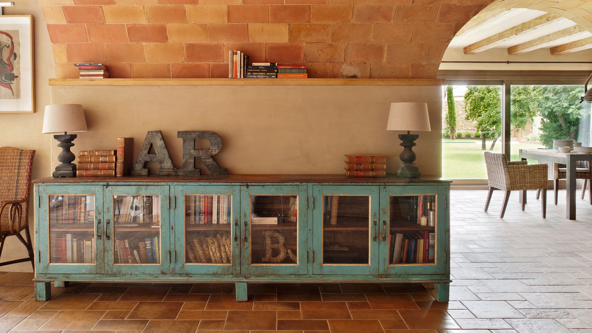 Recibidor con mueble bajo antiguo pintado en azul envejecido con puertas de cristal, lámparas de sobremesa, letras adornos, suelo y techo de barro