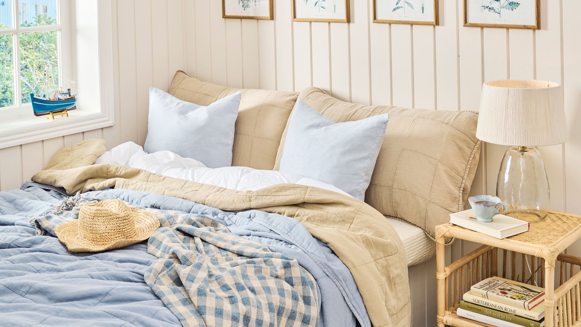 Dormitorio con lamas de madera blancas, cuadros, ropa de cama beige y azul, mesita de noche de ratán