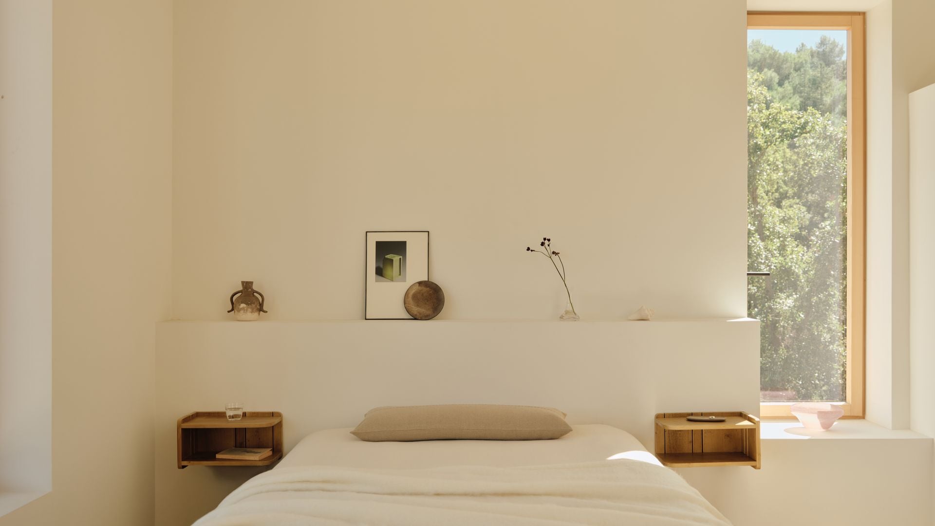 Dormitorio con cabecero de obra con repisa con adornos, mesitas de noche de madera suspendidas, ventana, ropa de cama blanca 