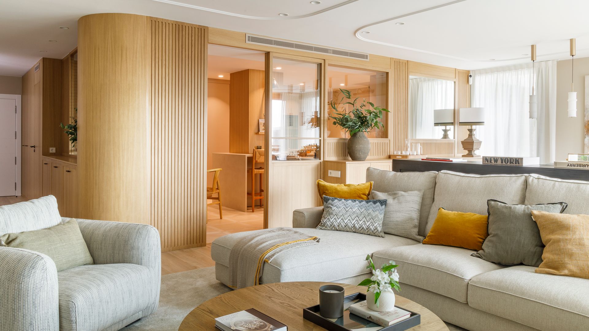 Salón comedor, abierto al recibidor, comunicado con la cocina con tabiques de cristal y madera, sofá blanco, mesa de centro redonda, cojines 