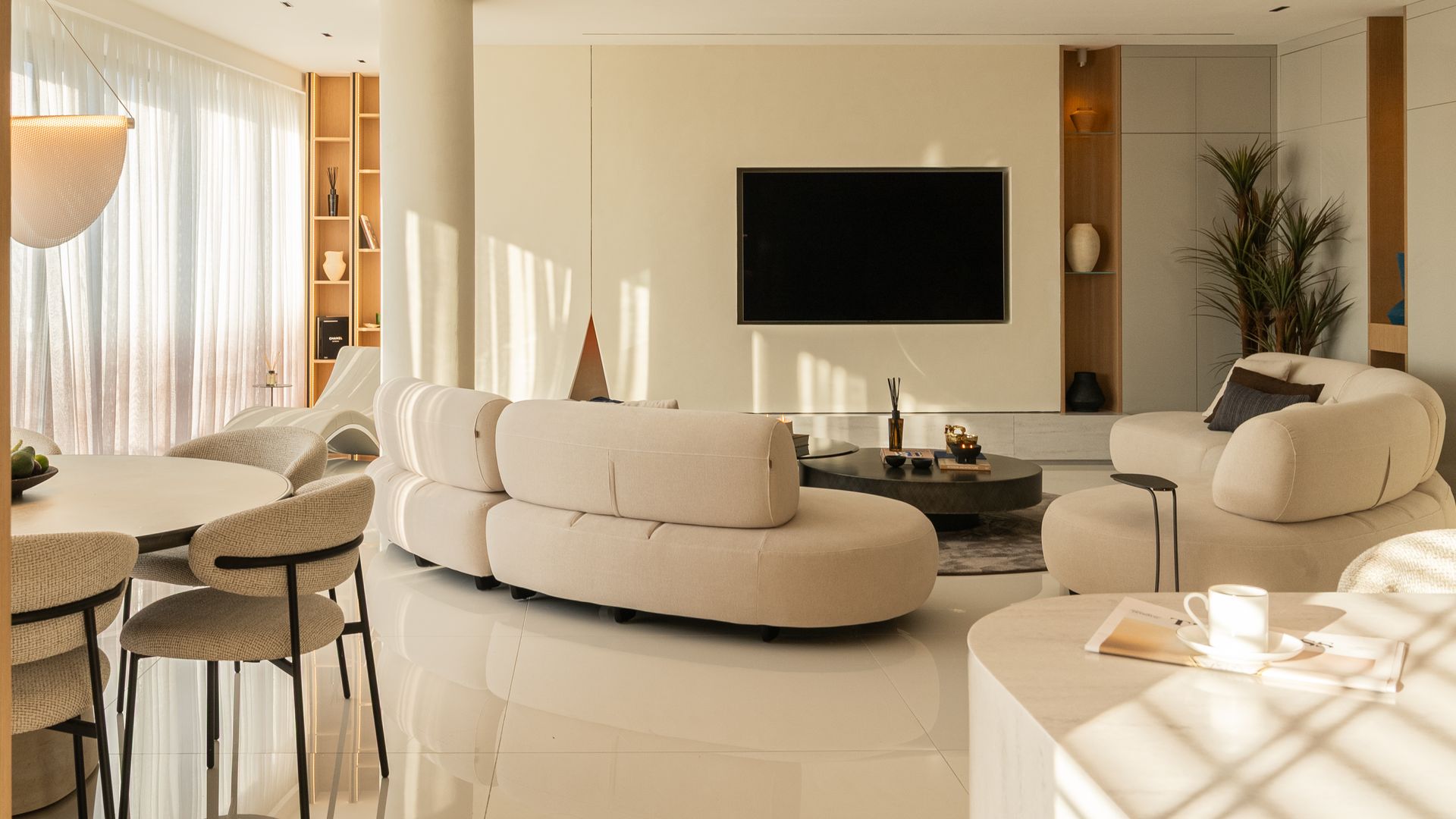 Salón comedor y cocina, televisión colgada en la pared, sofás curvos beiges con el respaldo bajo, mesa de comedor redonda, columnas, cortinas