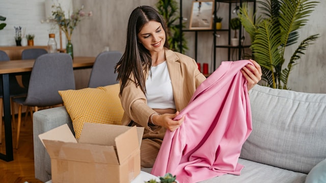 Mujer morena sentada en el sofá de su casa abriendo un paquete con una camisa rosa
