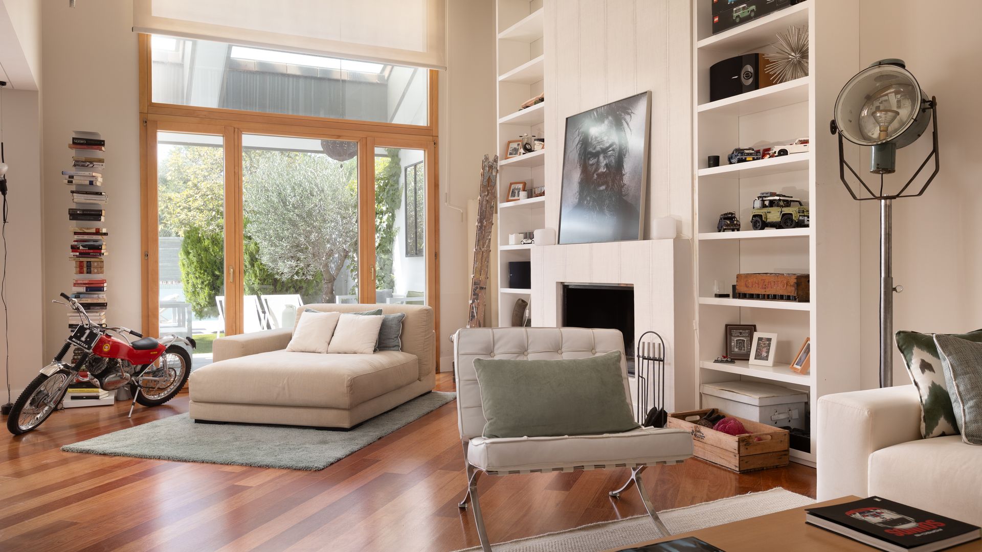 Salón con chimenea, librería blanca de suelo a techo, suelo0 de madera, oscura, silla Barcelona, chaise longue cama, fotografía en la repisa de la chimenea
