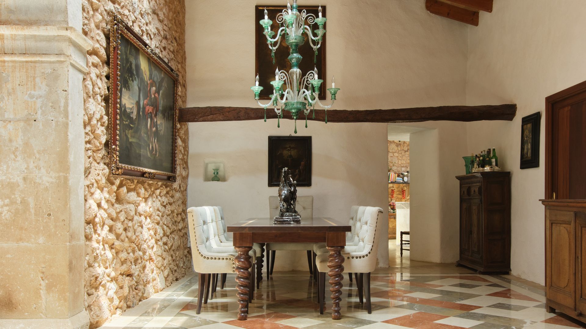 Comedor con techos altos, vigas de madera, lámpara de techo escultura, suelo de baldosas, mesa de comedor con patas torneadas, sillas de comedor tapizadas, cuadro en la pared