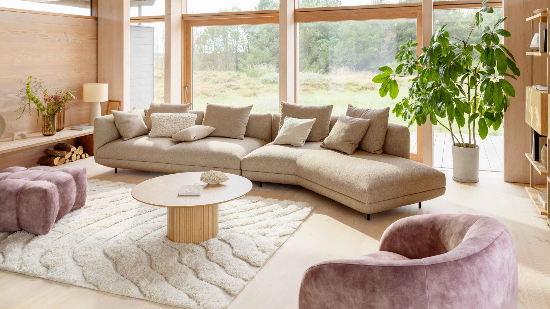 Salón con sofá modular beige, alfombra, mesa de centro redonda, sillones morados, planta alta en maceta, ventanales con perfiles de madera con salida al jardín
