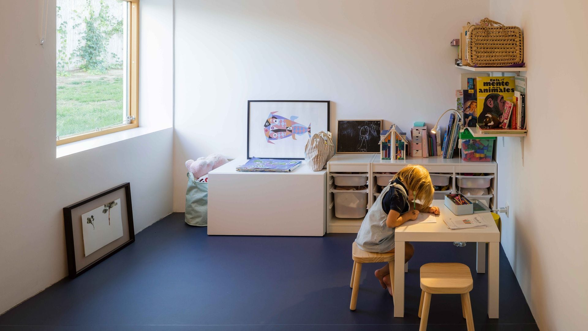 Habitación infantil con niña dibujando, suelo azul, mueble de almacenaje, mesa blanca, estanterías y cuadros