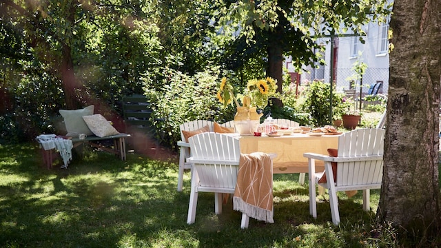 Mesa de jardín en verano con muchos árboles y césped. La mesa tiene un mantel crema sobre el que se despliega toda la vajilla y un jarrón con girasoles.