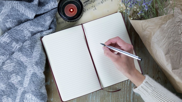 mujer escribiendo a mano en un cuaderno, con unas velas encendidas y unas flores