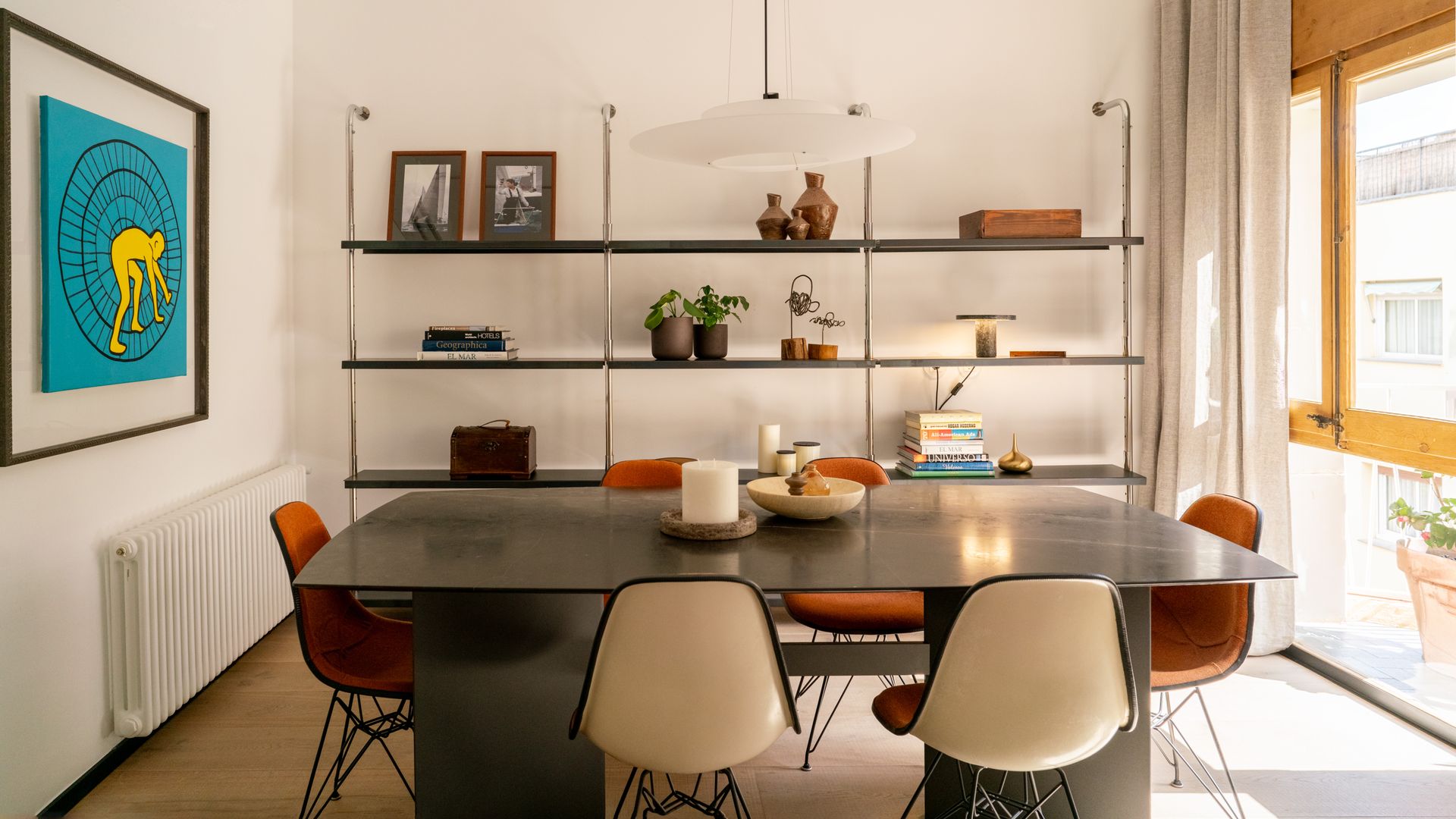 Comedor con gran mesa negra, ventanas con el marco de madera, sillas de estilo nórdico, estantería metálica con adornos y fotos, cuadro en la pared, radiador 