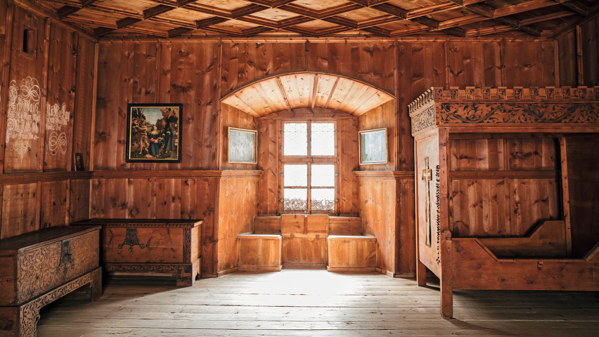 Habitación antigua de madera con muebles tallados, ventana arqueada y pintura religiosa.