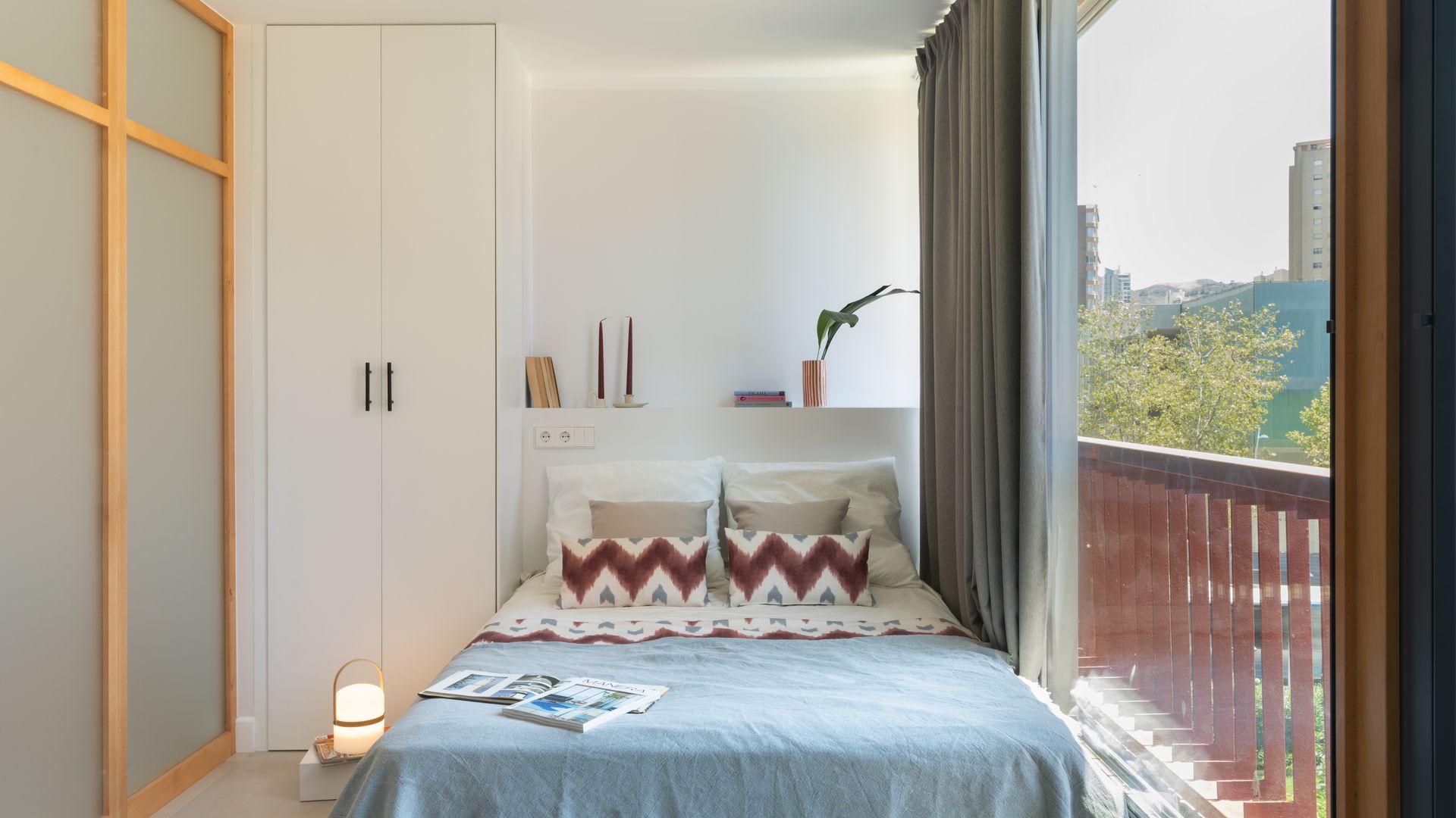 Dormitorio con gran ventanal, cabecero de obra con armario blanco, lámpara Cesta, cojines con motivos geométricos, plaid azul