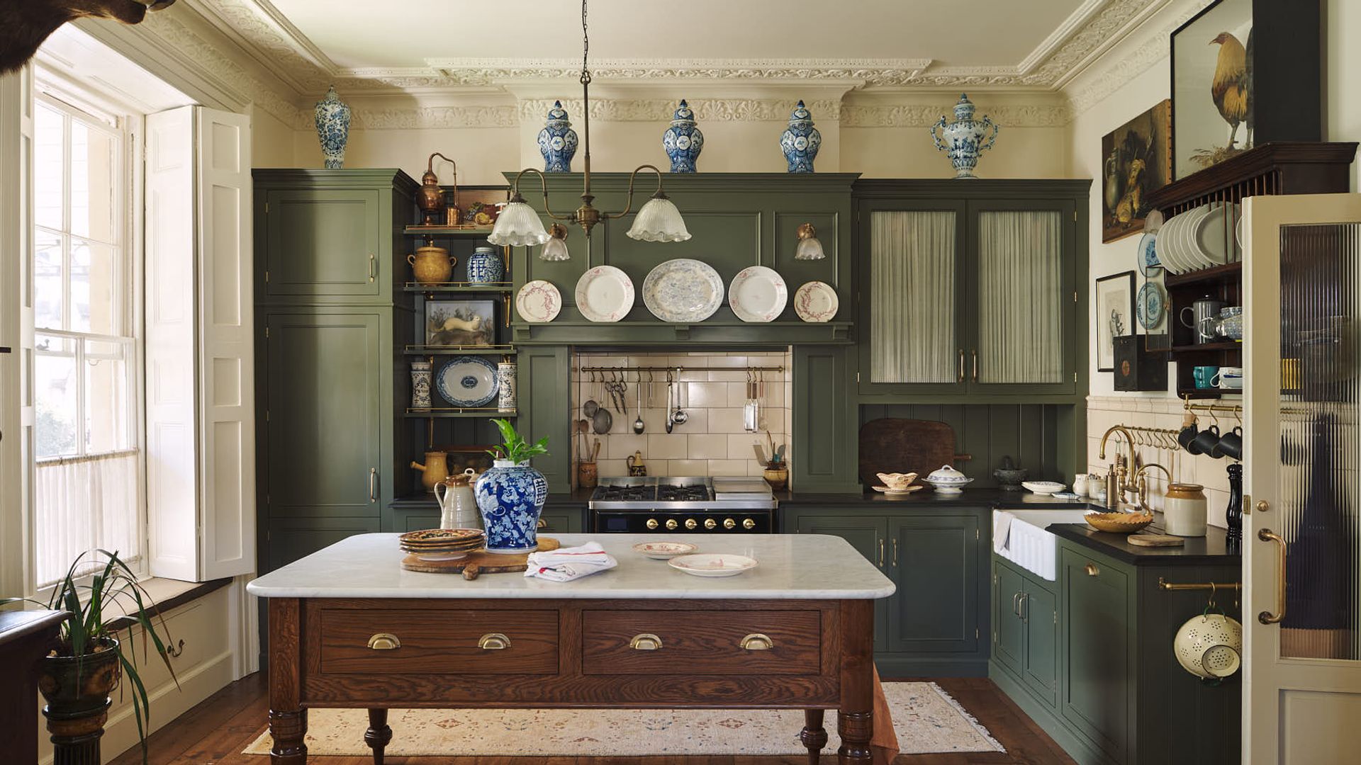 Cocina con muebles de madera verde, mesa tocinera en el centro, platos colgados en la pared, cortinillas en las vitrinas, jarrones en lo alto, suelo de madera oscura