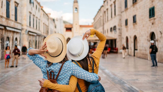 Dos mujeres amigas abrazándose haciendo turismo en su viaje por Dubrovnik, en Croacia, de espaldas, con sombreros y mochilas, en temporada de primavera