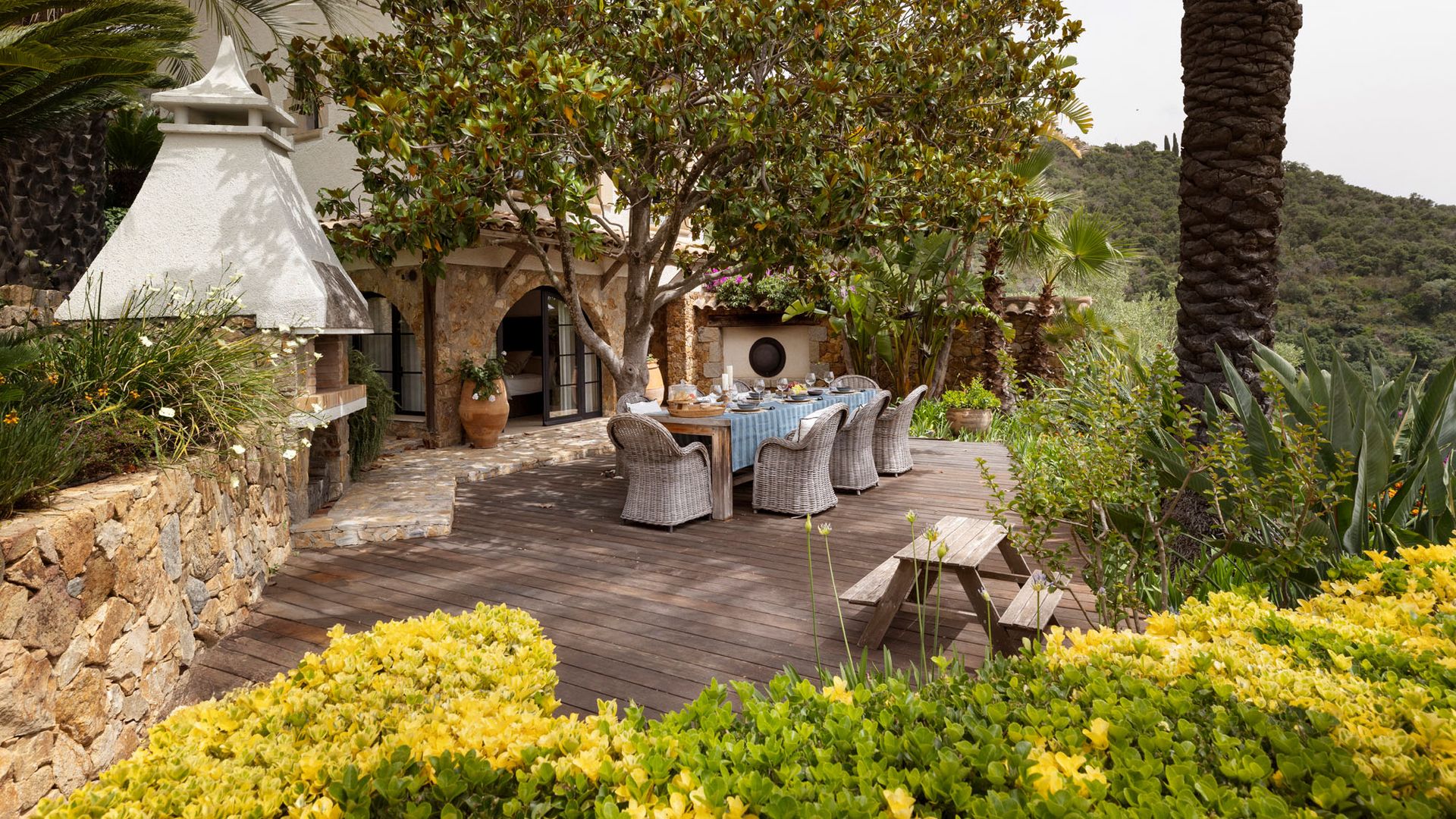 Jardín con comedor exterior con mesa XL con mantel, suelo de madera, sillas de fibras vegetales, horno y barbacoa, banco de madera, plantas
