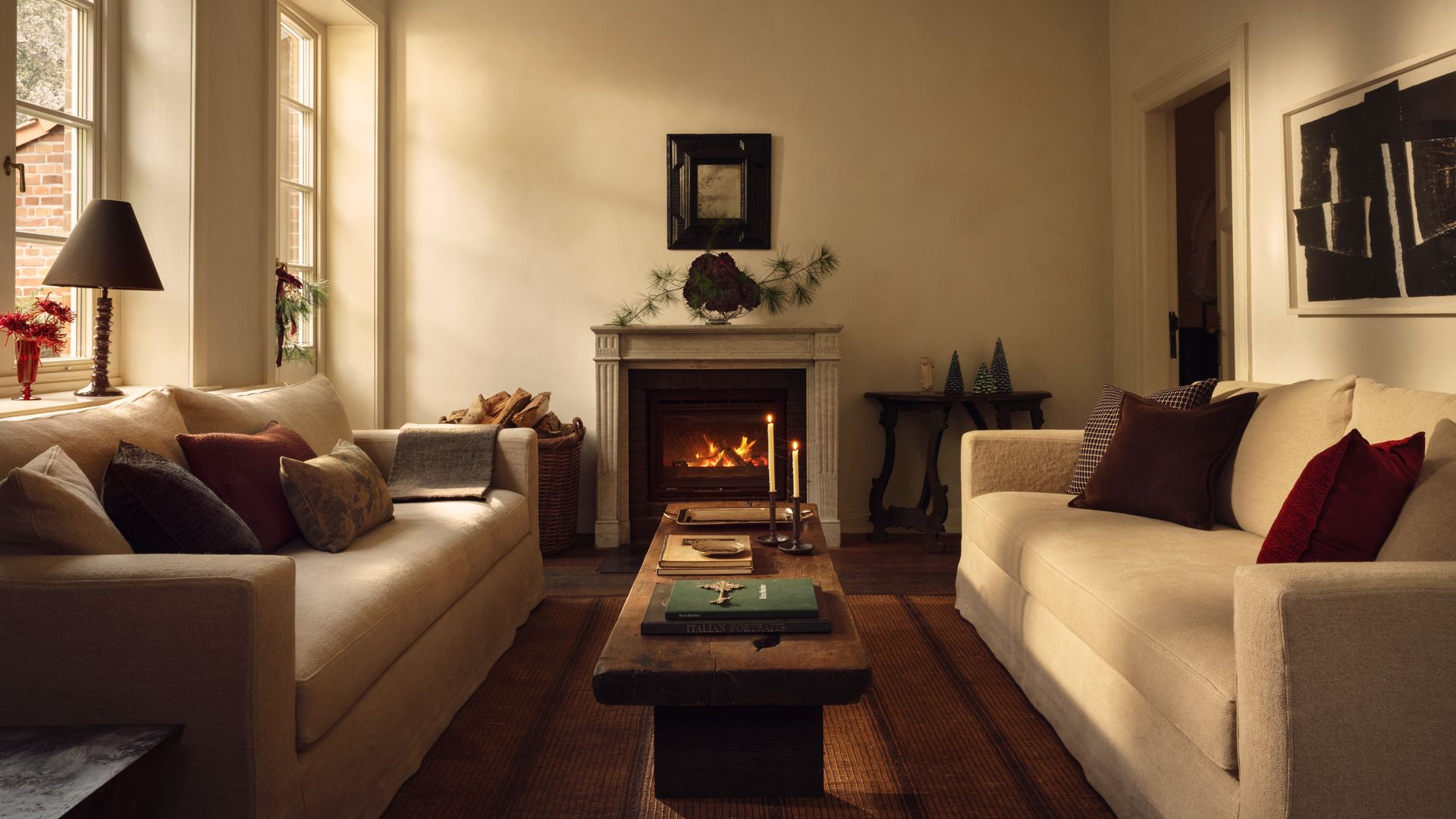 Salón con chimenea, ventanales, paredes en color crema y dos sofás enfrentados a juego, mesa de centro de madera, cojines color tierra y burdeos sobre los sofás, adornos navideños en el alféizar de la ventana y sobre la chimenea