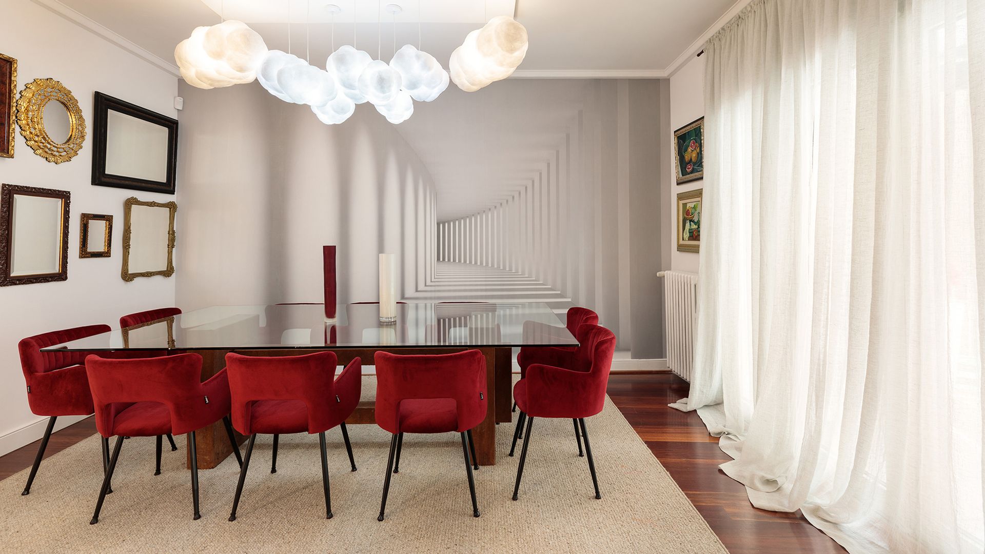 Comedor con mesa XL de cristal, alfombra, suelo de madera, sillas de comedor tapizadas en rojo, cuadros y marcos en las paredes, lámparas de techo blancas, cortinas blancas, papel pintado