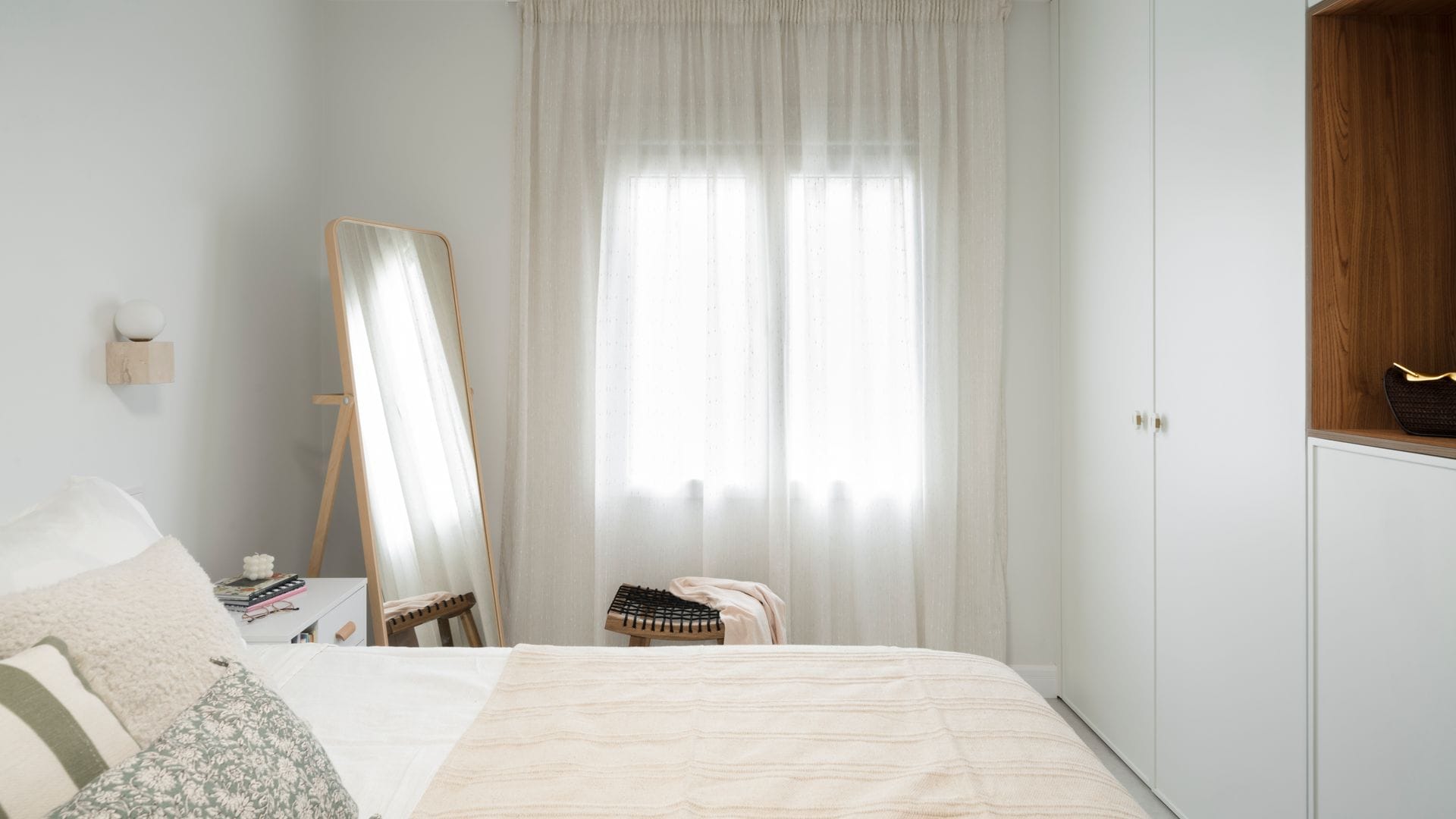 Dormitorio con armario a medida blanco con hueco abierto en madera, cortinas blancas, taburete, espejo de pie con el marco de madera, ropa de cama beige