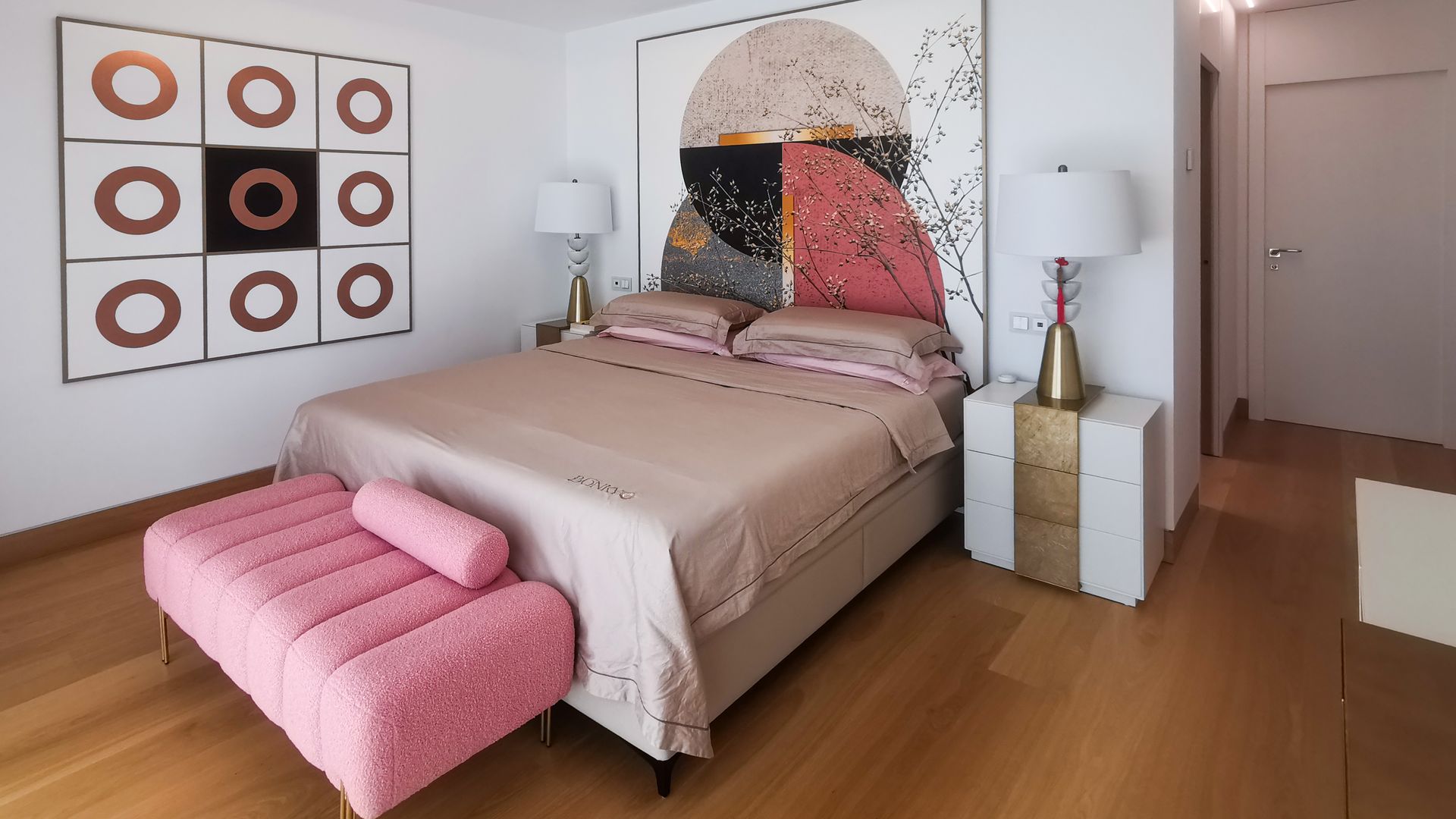 Dormitorio principal con cuadros en la pared del cabecero, banqueta rosa, mesita de noche dorada y blanca, suelo de madera, plafón en el techo