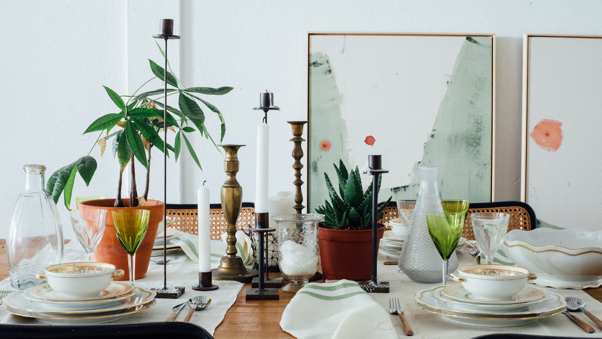 Mesa de comedor con manteles individuales, candelabros, vajilla blanca con el borde dorado, plantas, copas verdes 