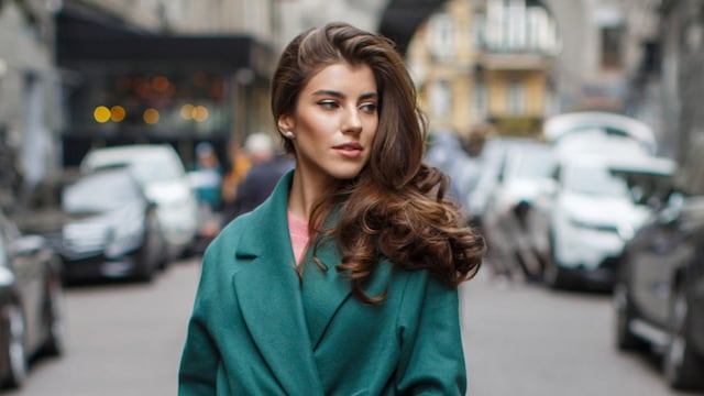 Mujer en la calle con pantalones blancos, abrigo y bolso verdes y pelo ondulado con volumen