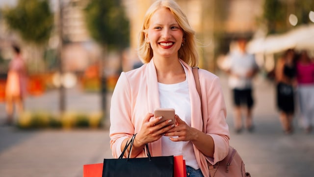 Mujer rubia sonriente en la calle con bolsas de compras y teléfono móvil en la mano