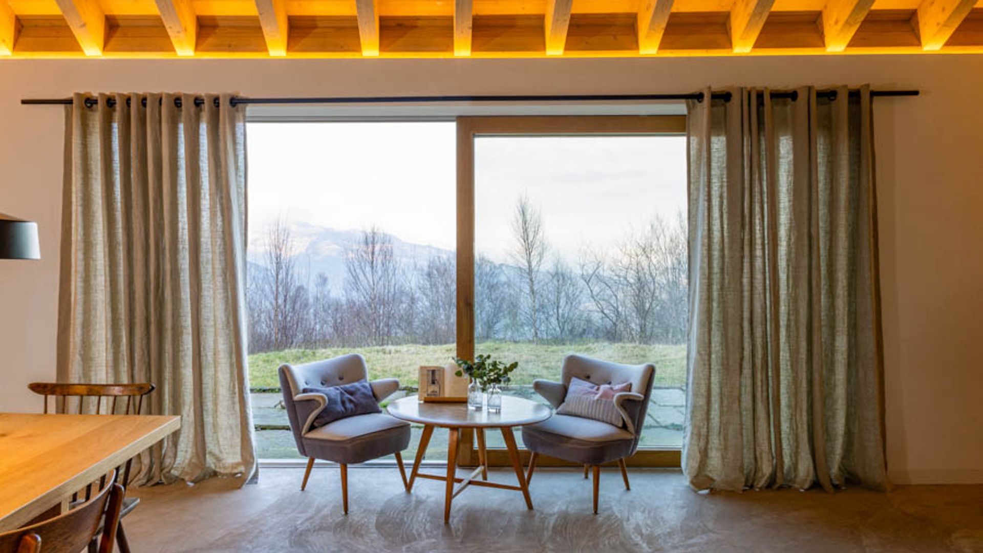 Salón comedor con mesa de madera, ventanal con cortinas, rincón con mesa y butacas, techo de madera
