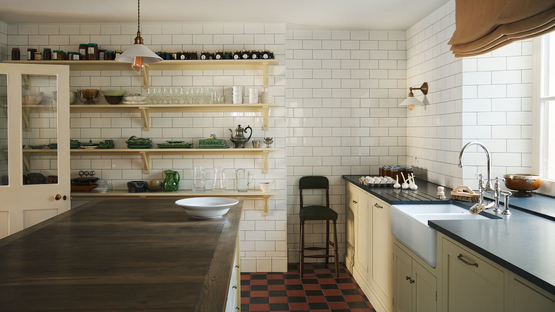 Cocina vintage con suelo de damero rojo y negro, muebles clásicos de color blanco mantequilla, azulejos blancos brillantes, tipo metro, baldas de madera con botes y vajilla, silla alta, estor