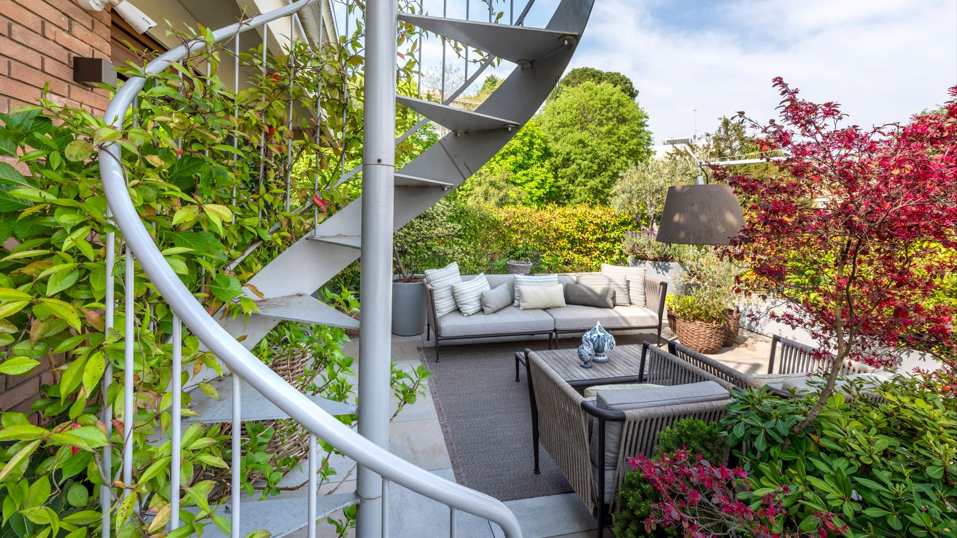Terraza con plantas y enredaderas, escalera de caracol, mesa y sofás grises, lámpara de pie