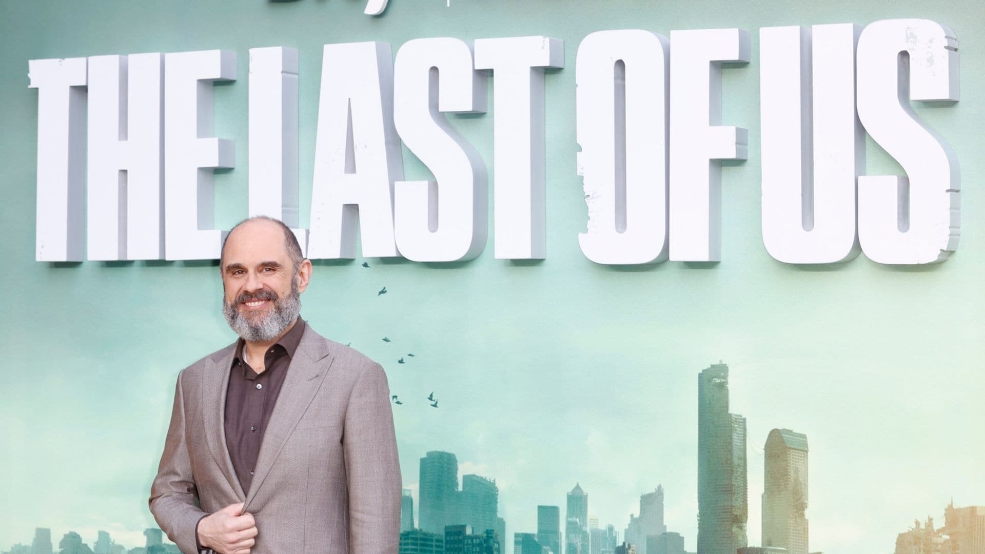 Craig Mazin sonríe frente a un cartel de 'The Last of Us' con ciudad en el fondo.
