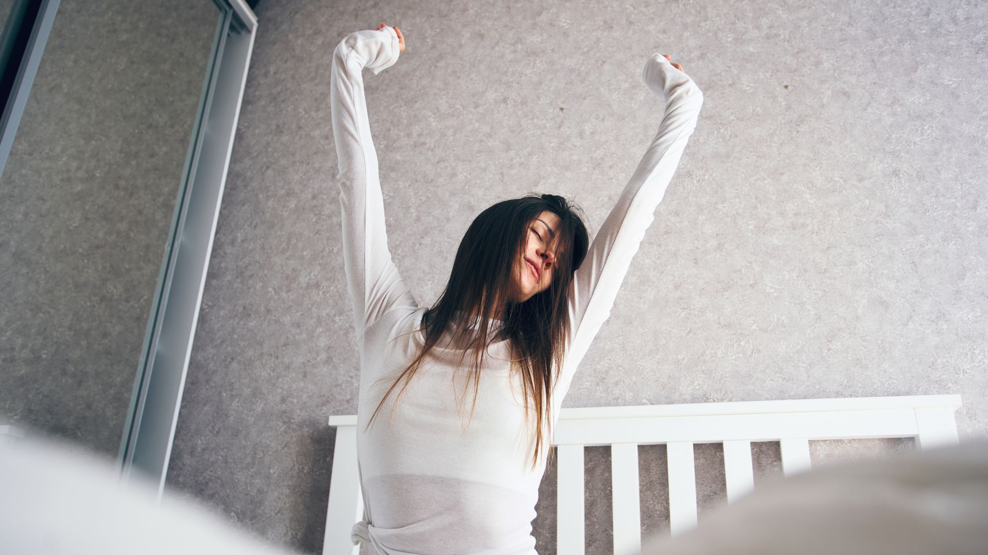 Mujer joven despertándose en una cama blanca, con un pijama todo blanco y el pelo suelto, estirándose hacia arriba con los ojos cerrados.