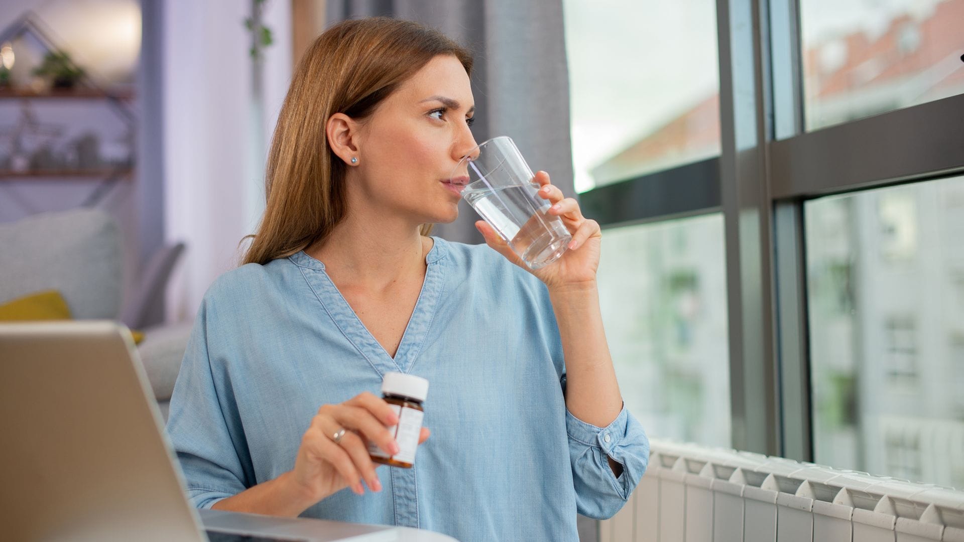 mujer tomando unas pastillas con un vaso de agua, mientras mira por la ventana