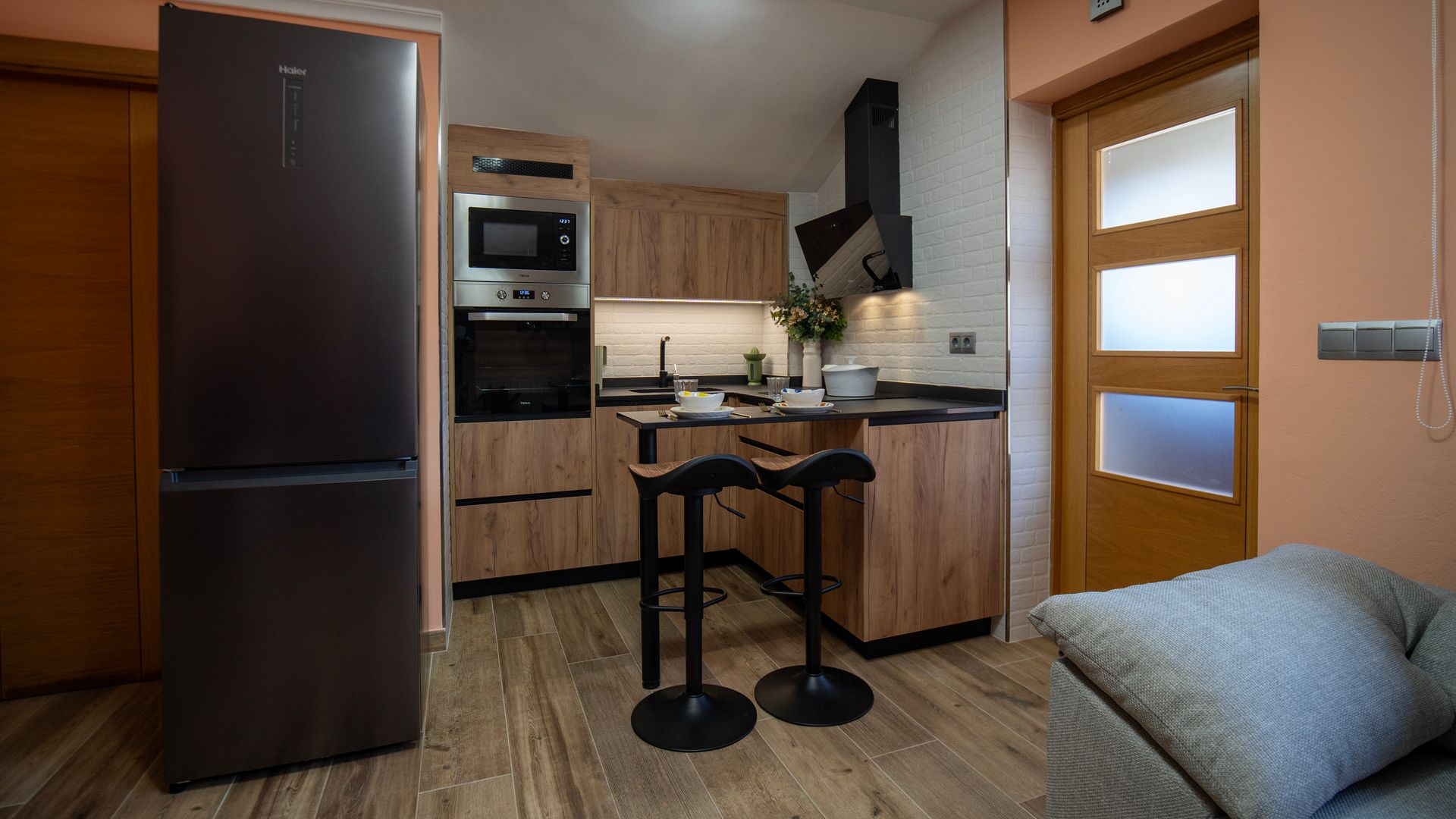 Cocina abierta con barra, frigorífica negro, muebles y suelo de madera, encimera negra