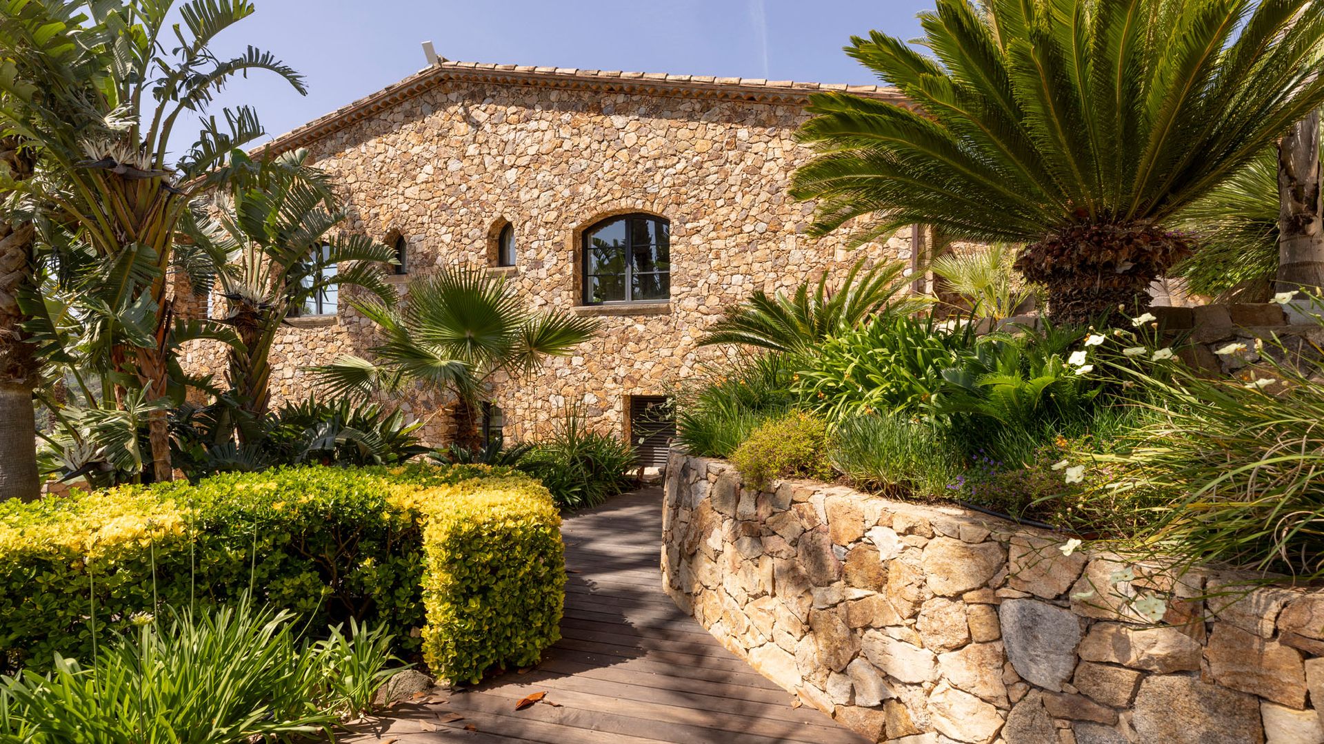 Masía de piedra con muro de piedra, palmeras y vegetación 