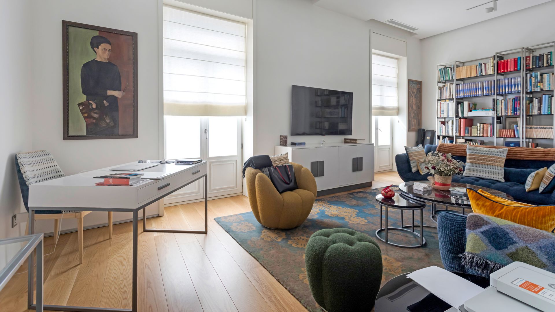 Salón con zona de escritorio, mueble de televisión, sofás y sillones de color azul y mostaza, librería llena de libros, alfombra 