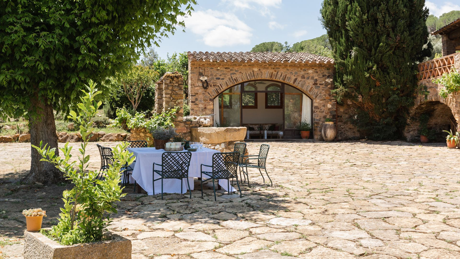 Jardín con mesa de verano, suelo de piedra, mantel blanco, sillas negras y velas
