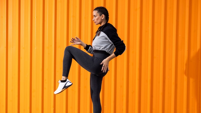 Mujer joven con leggins grises y chaqueta de deporte, con una pierna levantada sobre fondo naranja
