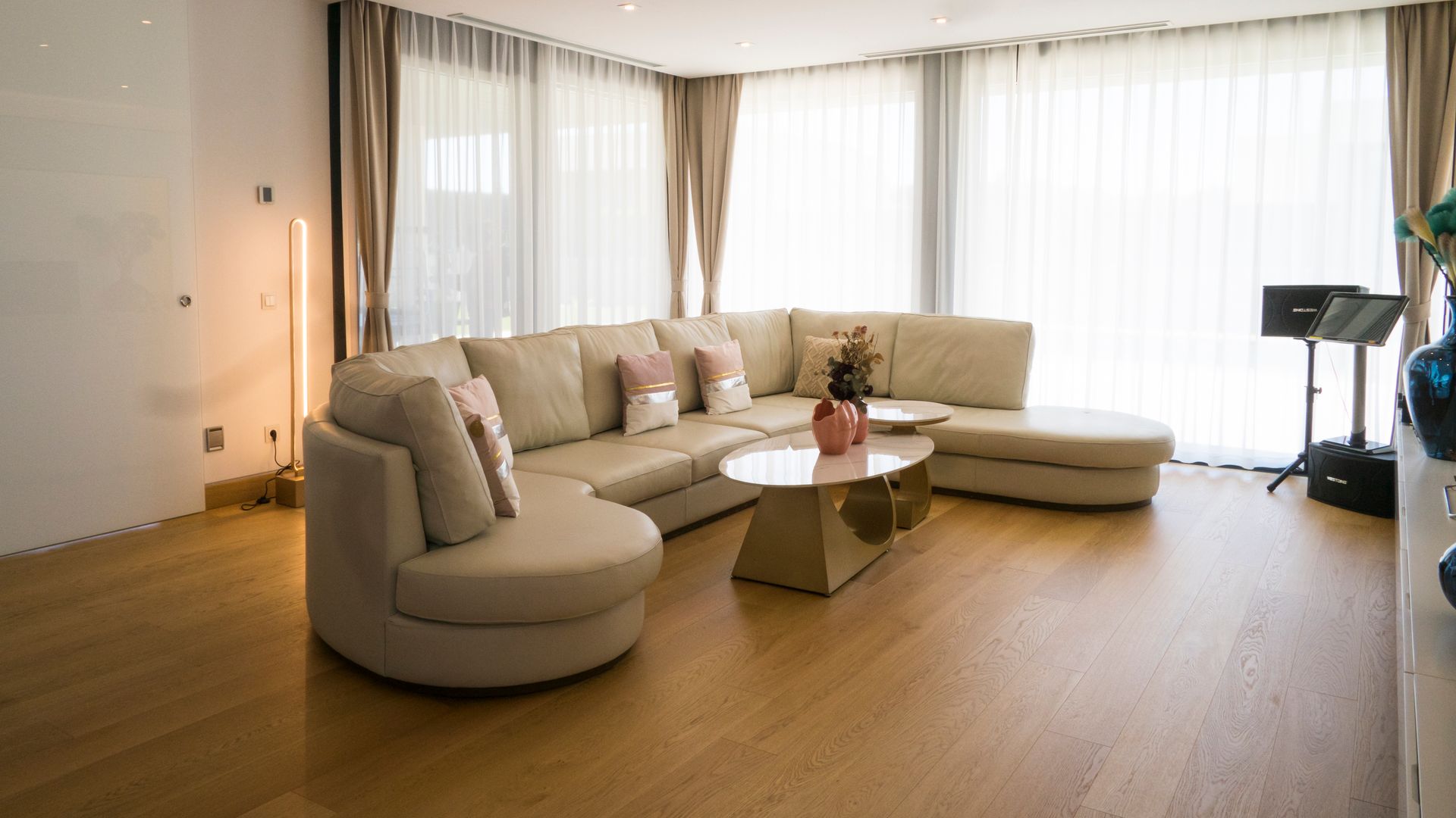 Salón con suelo de madera, cortinas blancas y marrones, sofá de cuero blanco, mesa de centro, focos empotrados en el techo, puerta corredera de cristal opaco